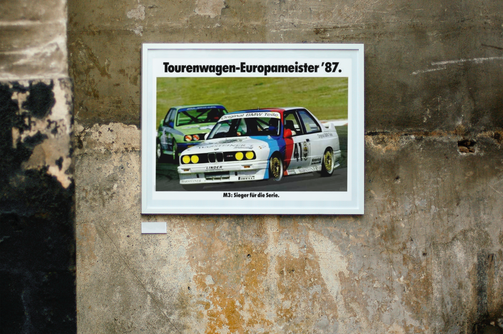 BMW E30 M3 shown in motion, racing on a track. Title above the photo reads European Touring Car Champion 1987. The frame is displayed on a beautiful concrete wall.