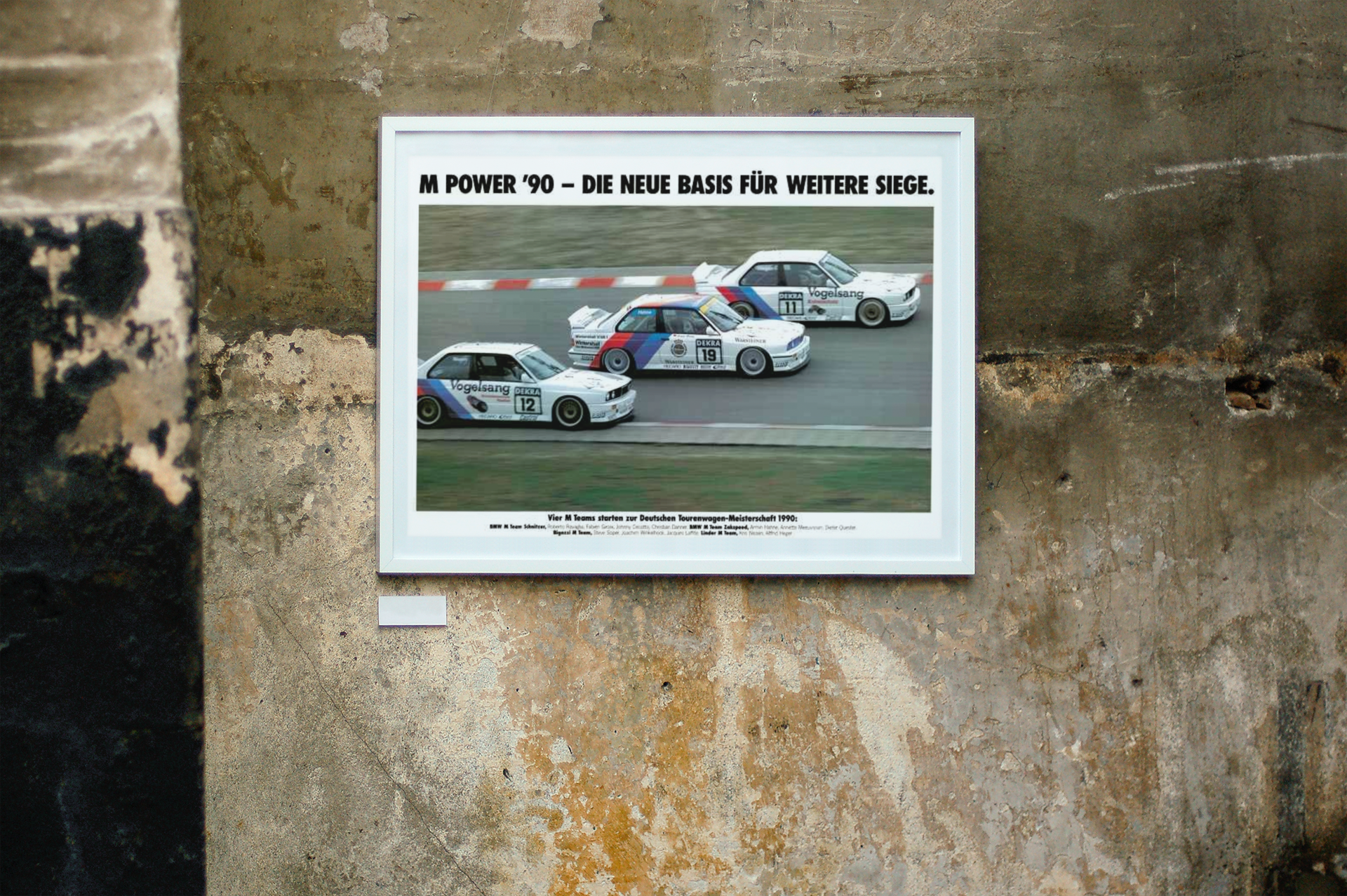 Three BMW E30 M3 race cars shown in motion, racing on a track. Title above the photo reads M Power '90 - The new basis for further victories. The frame is displayed on a beautiful concrete wall.